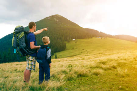 Father and son with backpacks hiking together in summer mountains. Back view of dad and child...