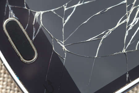 Close-up detail of black old cellphone with cracked screen on light cloth background. Gadget repair...