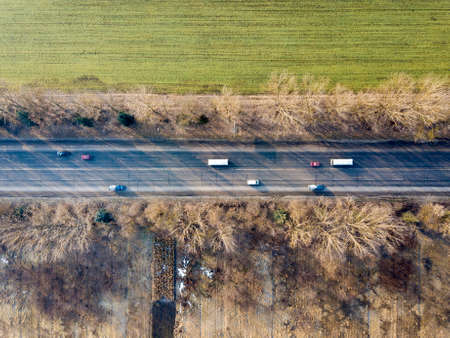 Aerial view of straight road with moving cars, trees and green fields on sunny day. Drone...