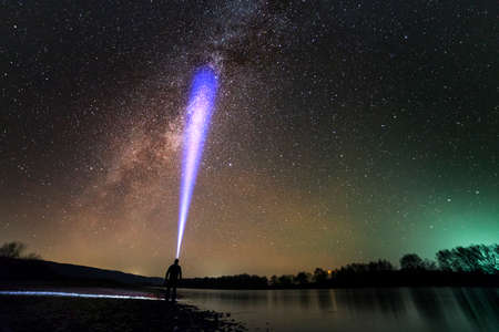 Back view of man with head flashlight standing on river bank, long blue beam across beautiful dark starry sky. Night photography concept. Wide panorama, copy space background.の写真素材