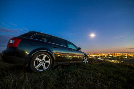 Black car parked at night in green meadows on copy space background of lights of distant city buildings and bright blue sky with first star at sunset.の写真素材