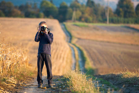 Young child boy with photo camera taking picture of wheat field on blurred rural background.