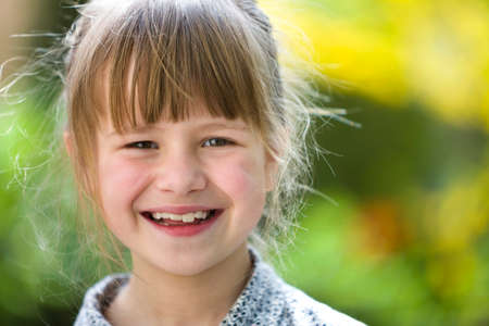 Cute pretty child girl with gray eyes and fair hair smiling in camera outdoors on blurred sunny green and yellow bright bokeh background.の写真素材