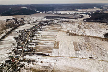 Top view of empty snowy fields, houses along road and woody hills on blue sky background. Aerial...