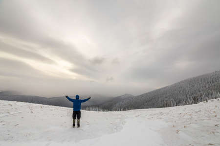 Back view of tourist hiker in warm clothing with backpack standing with raised arms on clearing covered with snow on spruce forest mountain and cloudy sky   .の写真素材