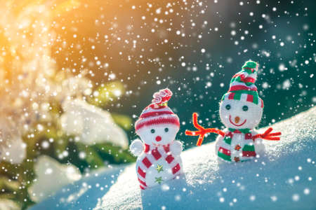 Two small funny toys baby snowman in knitted hats and scarves in deep snow outdoors near pine tree branch. Happy New Year and Merry Christmas greeting card.の写真素材