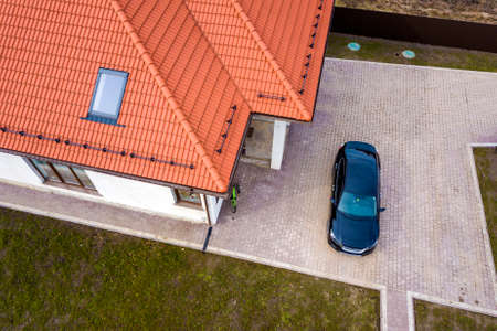 Aerial top view of house metal shingle roof with attic windows and black car on paved yard.