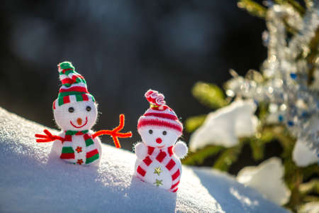 Two small funny toys baby snowman in knitted hats and scarves in deep snow outdoors near pine tree branch. Happy New Year and Merry Christmas greeting card.の写真素材