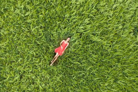View from above of young attractive slim woman in red dress laying with closed eyes in green wheat field.の写真素材