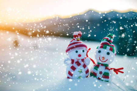 Two small funny toys baby snowman in knitted hats and scarves in deep snow outdoors on blurred mountains landscape background. Happy New Year and Merry Christmas greeting card theme.の写真素材