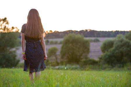 Young woman standing alone on a field with green grass enjoying warm sunset.の写真素材