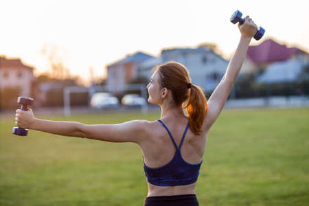 Portrait of athletic teenage girl in fitness wear exercising with blue dumbbells outdoors in park....