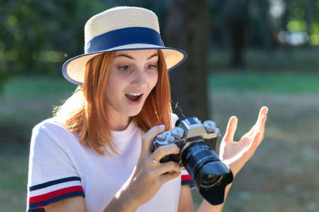 Pretty teenage girl with red hair looking amazed in photo camera in summer park.の写真素材