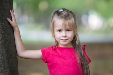 Portrait of a pretty little child girl standing near big tree trunk in summer park outdoors.の写真素材