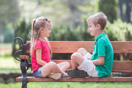 Portrait of two pretty cute children boy and girl having fun time on a bench in summer park outdoors.の写真素材