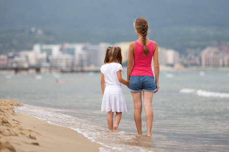 Mother and daughter girl walking together on sand beach in sea water in summer with bare feet in warm ocean waves.の写真素材