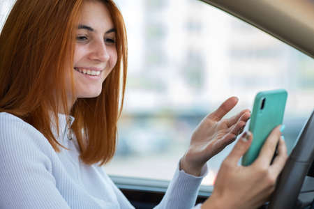 Happy redhead girl texting on her mobile phone behind the wheel driving a car.の写真素材
