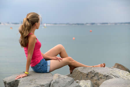 Young woman in summer clothes sitting on big boulders on sea shore looking on horizon.
