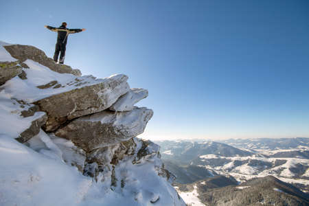 Silhouette of alone tourist standing on snowy mountain top in winner pose with raised hands enjoying view and achievement on bright sunny winter day. Adventure, outdoors activities, healthy lifestyle.の写真素材