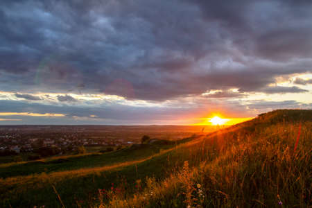 Sunset in dramatic cloudscape over wild herb field with distant city in view.