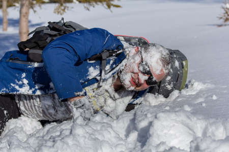 Hiker in sunglasses with backpack lay down with snow on his face in winter.