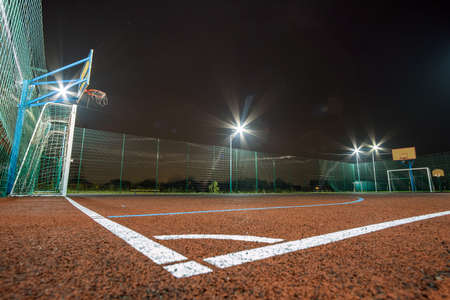 Outdoors mini football and basketball court with ball gate and basket surrounded with high protective fence brightly illuminated with spotlight lamps at night.の写真素材