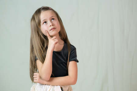 Close-up portrait of happy smiling little girl with long hair holding finger to her cheek thinking.
