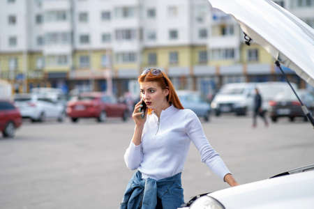 Young woman standing near broken car with popped hood talking on her mobile phone while waiting for...