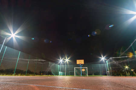 Outdoors mini football and basketball court with ball gate and basket surrounded with high protective fence brightly illuminated with spotlight lamps at night.の写真素材