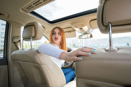 Wide angle view of young redhead woman driver driving a car backwards looking behind.