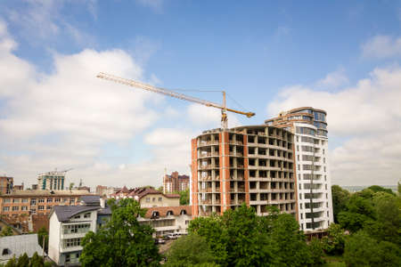 Apartment or office tall building unfinished under construction among green tree tops. Tower cranes on bright blue sky copy space background.の写真素材