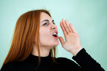 Pretty redhead girl shouting loudly holding her palm to face.