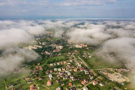 Aerial view of white clouds above a town or village with rows of buildings and curvy streets between green fields in summer. Countryside landscape from above.の写真素材