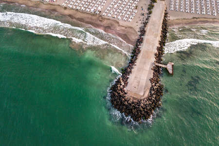 Aerial view of a concrete pier on sea shore with wave breaking stones.