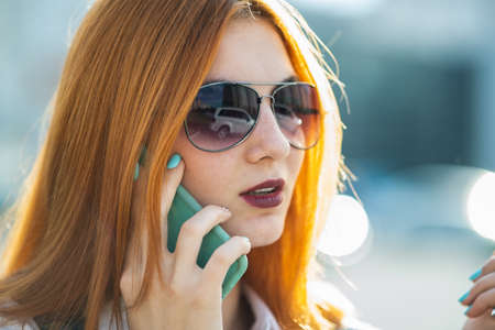 Closeup portrait of fashionable young redhead woman in sunglasses talking on cell phone outdoors in...
