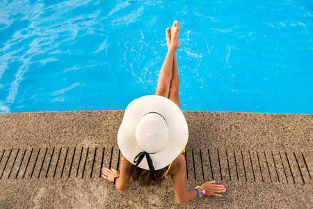 Top down view of young woman wearing yellow straw hat resting near swimming pool with clear blue...