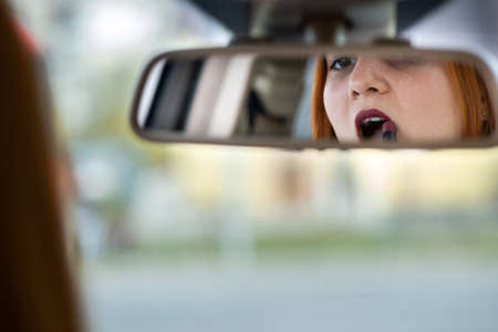 Closeup of a young redhead woman driver correcting her makeup with dark red lipstick looking in car rearview mirror behind steering wheel of a vehicle.の写真素材