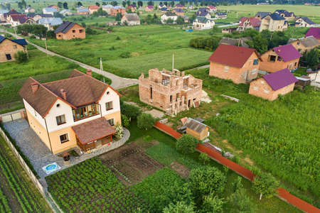 Aerial top view of a private house with attic windows on roof, paved yard with green grass lawn and building site with concrete foundation floor and stacks of yellow bricks for construction..の写真素材