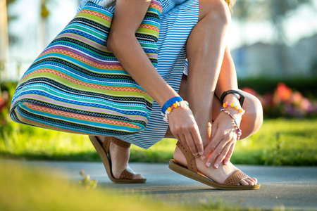 Close up of woman hands tying her open summer sandals shoes on sidewalk in sunny weather.