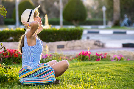 Young woman wearing light blue summer dress and yellow straw hat relaxing on green grass lawn in summer park. Girl in casual outfit resting outdoors enjoying free time in warm morning.の写真素材