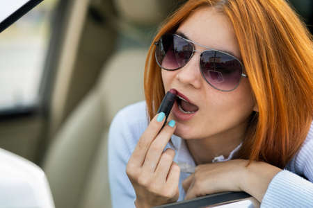 Closeup of a young redhead woman driver correcting her makeup with dark red lipstick looking in car rearview mirror behind steering wheel of a vehicle.の写真素材