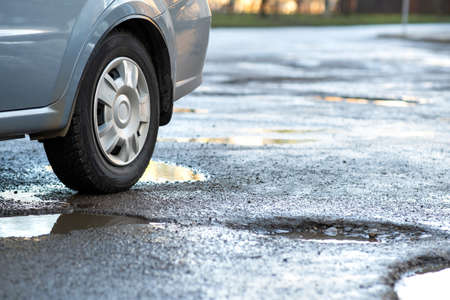Close up of car wheel on a road in very bad condition with big potholes full of dirty rain water pools.の写真素材
