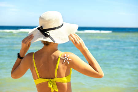Back view of young woman tanning at the beach with sunscreen cream in sun shape on her shoulder. UV...