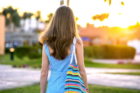 Young woman wearing light blue summer dress holding fashionable shoulder bag standing outside enjoying warm weather in summer park at sunset.の写真素材