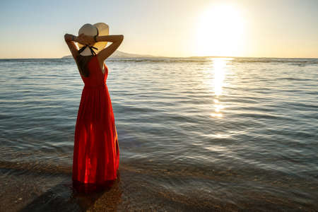 Young woman wearing long red dress and straw hat standing in sea water at the beach enjoying view of rising sun in early summer morning.の写真素材