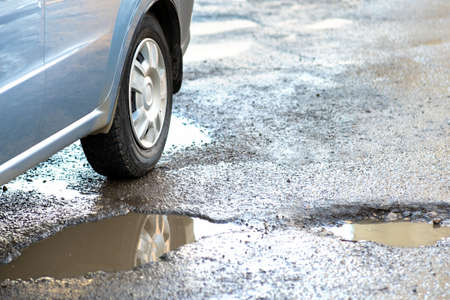 Close up of car wheel on a road in very bad condition with big potholes full of dirty rain water pools.の写真素材
