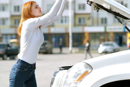 Young stressed woman driver near broken car with popped hood having a prbreakdown problem with her...