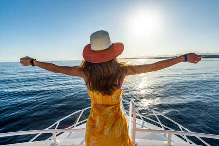 Young woman with long hair wearing yellow dress and straw hat standing with raised hands on white yacht deck enjoying view of blue sea water.の写真素材