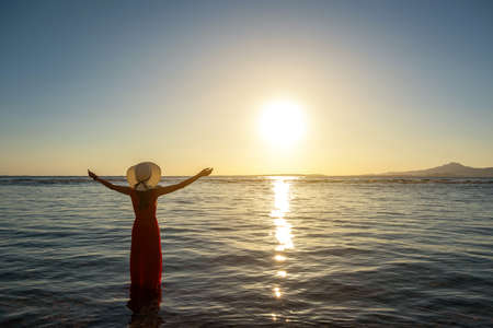 Young woman wearing long red dress and straw hat raising hands standing in sea water at the beach...