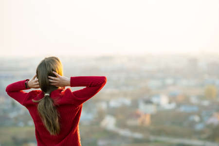 Young woman in red jacket standing with her hands behind head outdoors enjoying evening view. Relaxing, freedom and wellness concept.の写真素材